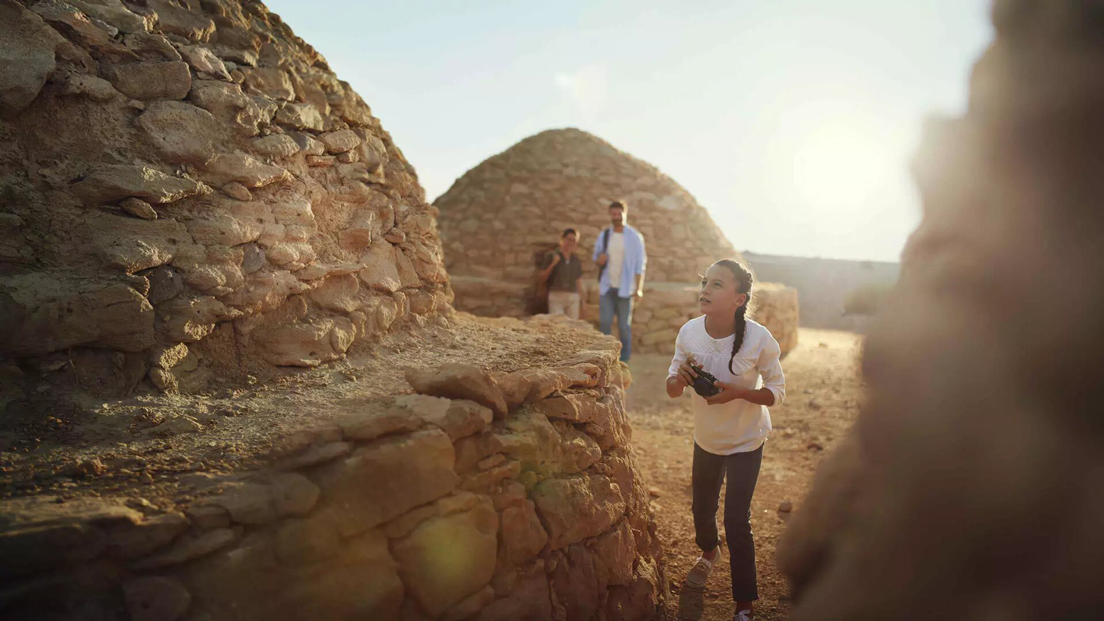 A woman wearing white shirt and black pants holding a camera, walking on rocky ground with a rock wall on the left. Two people stand in the
