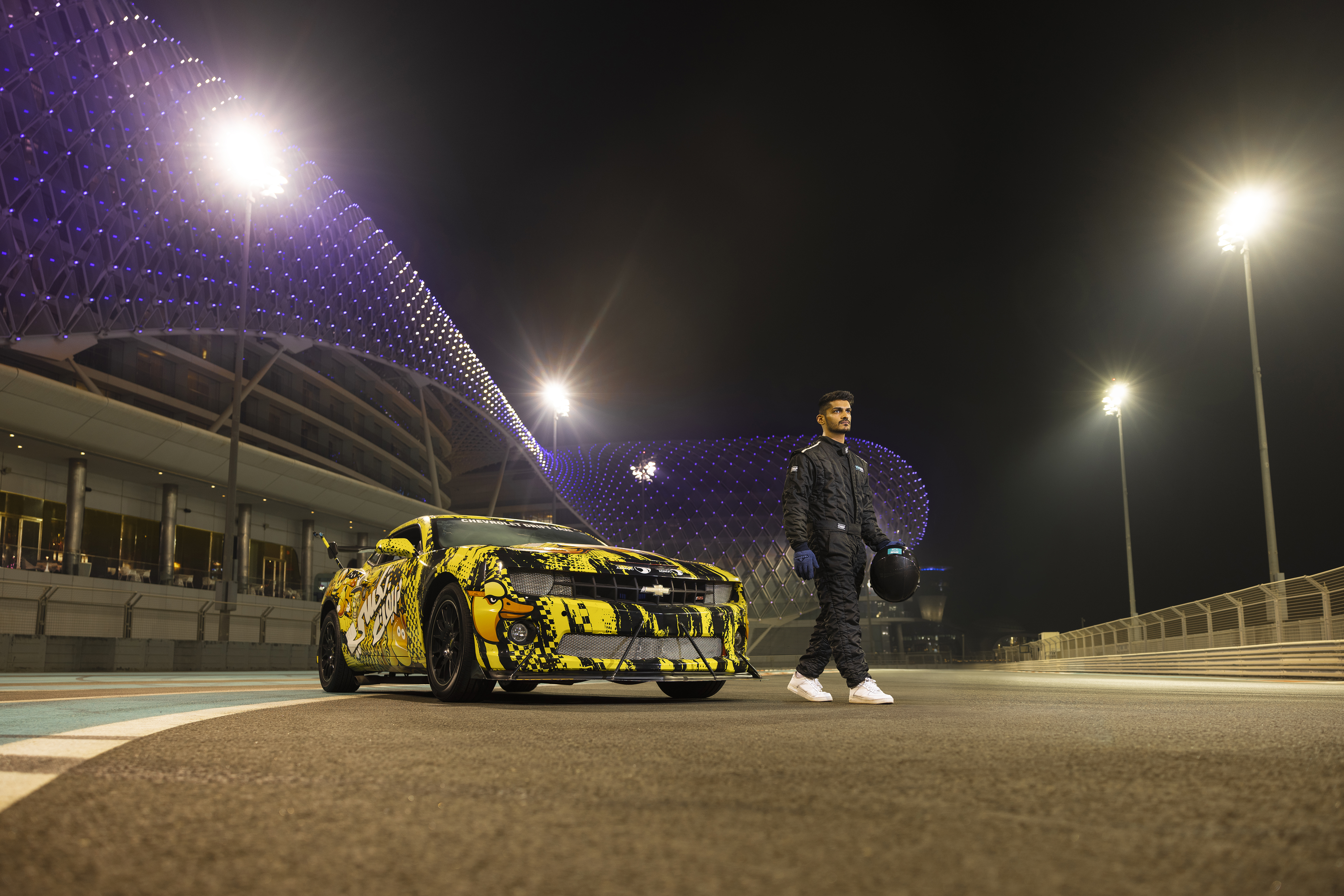 Race car driver walking past custom sports car at Yas Marina Circuit in Abu Dhabi under night lights.