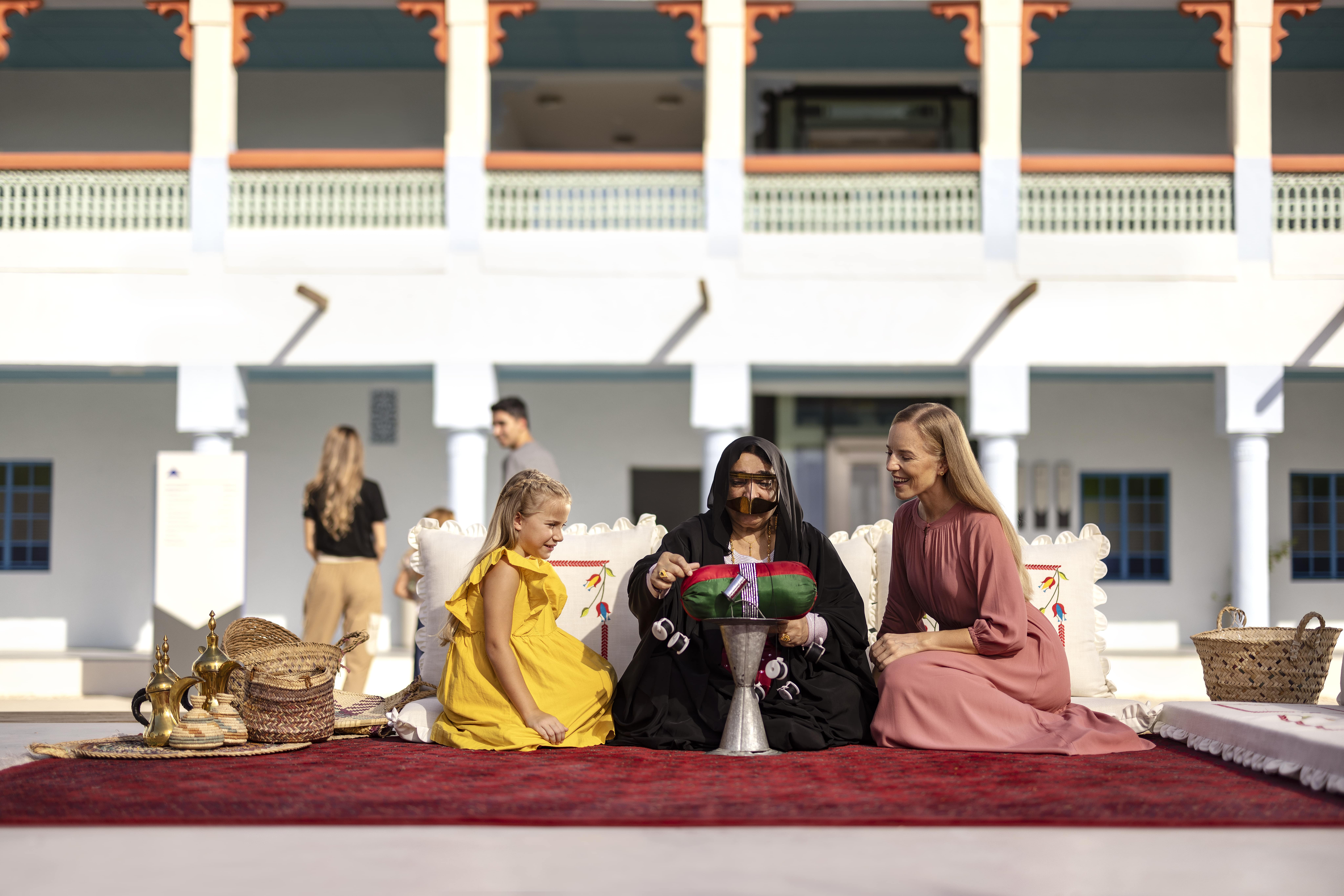 Traditional Emirati woman hosting guests with coffee and baskets in a cultural courtyard setting.