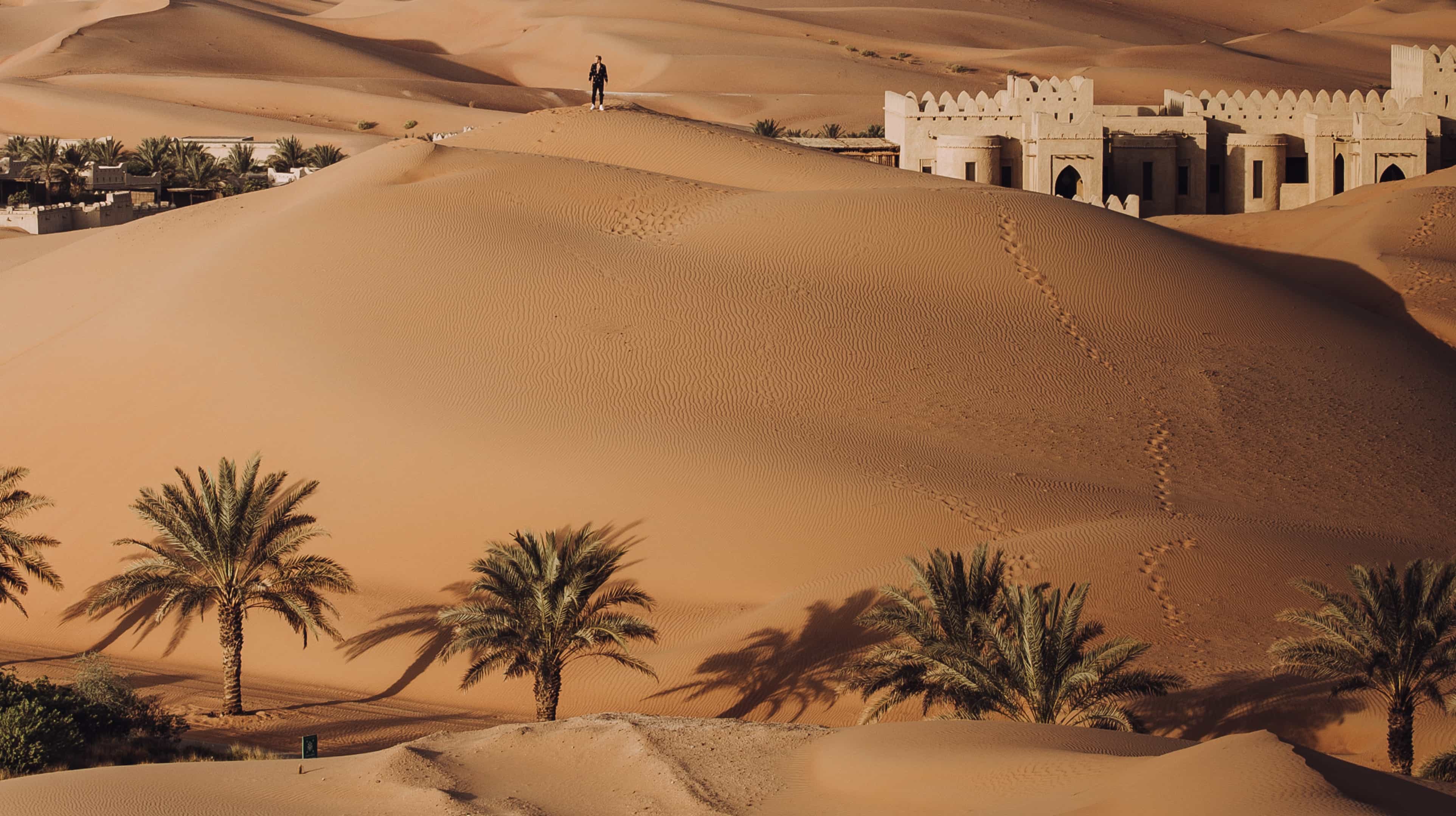 Vista dall'alto del deserto Empty Quarter con l' Anantara Resort, palme e un uomo che cammina sulle dune segnate da orme