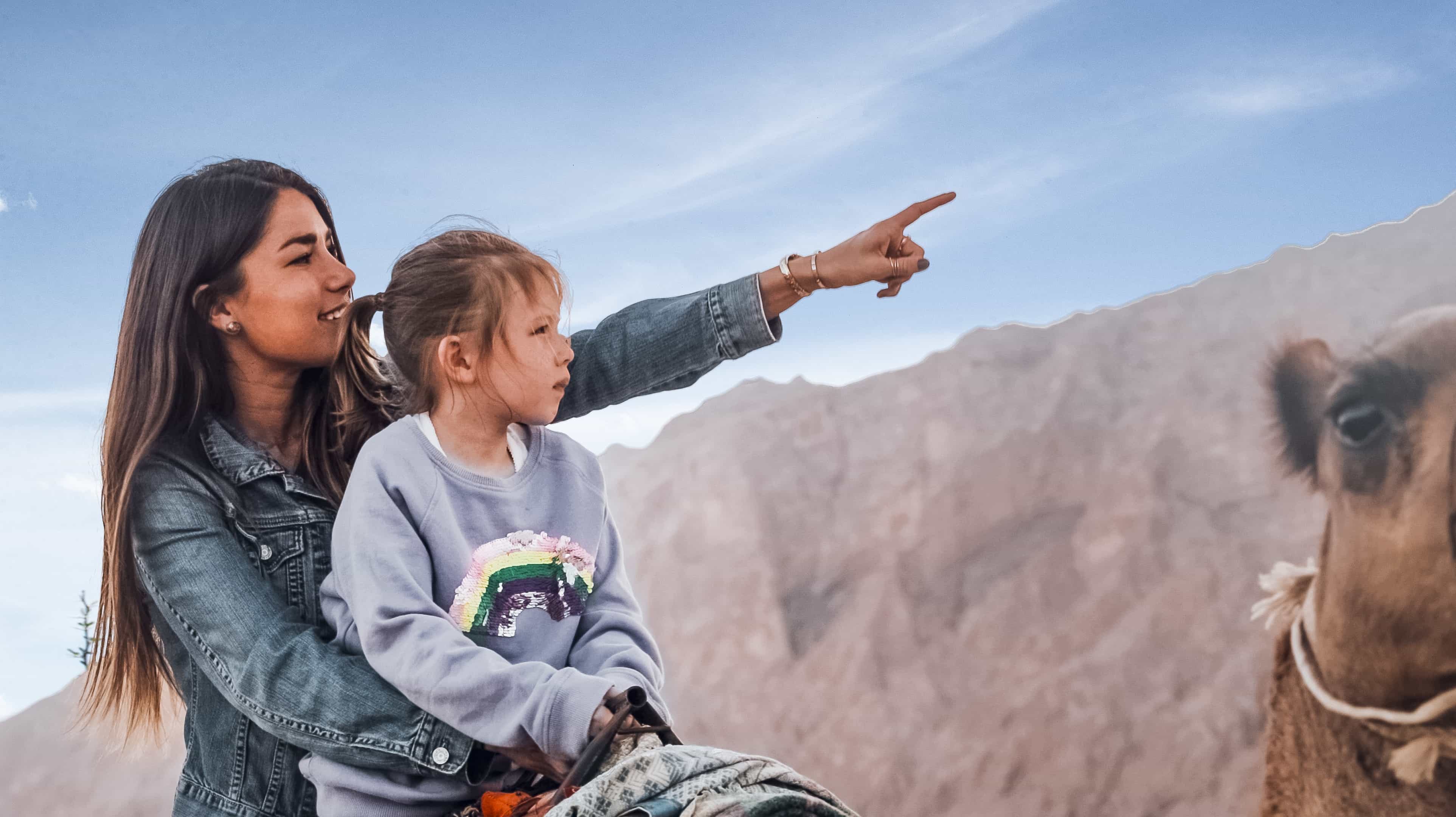 Woman and girl on a camel's back pointing at Jebel Hafit