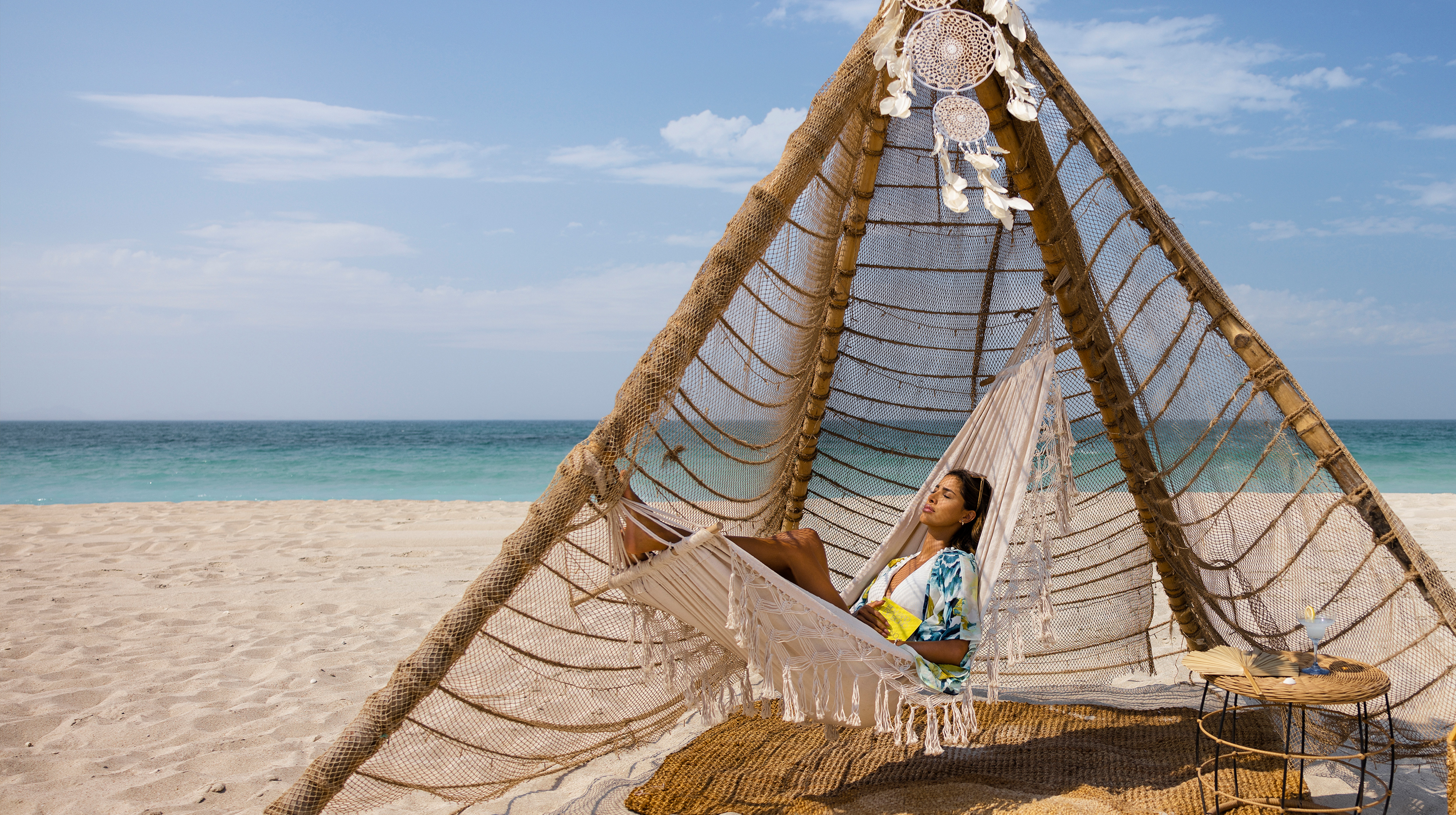 Woman relaxing on a hammock under a bamboo shelter at Kai Beach, Abu Dhabi, with the ocean and sandy beach in the background.