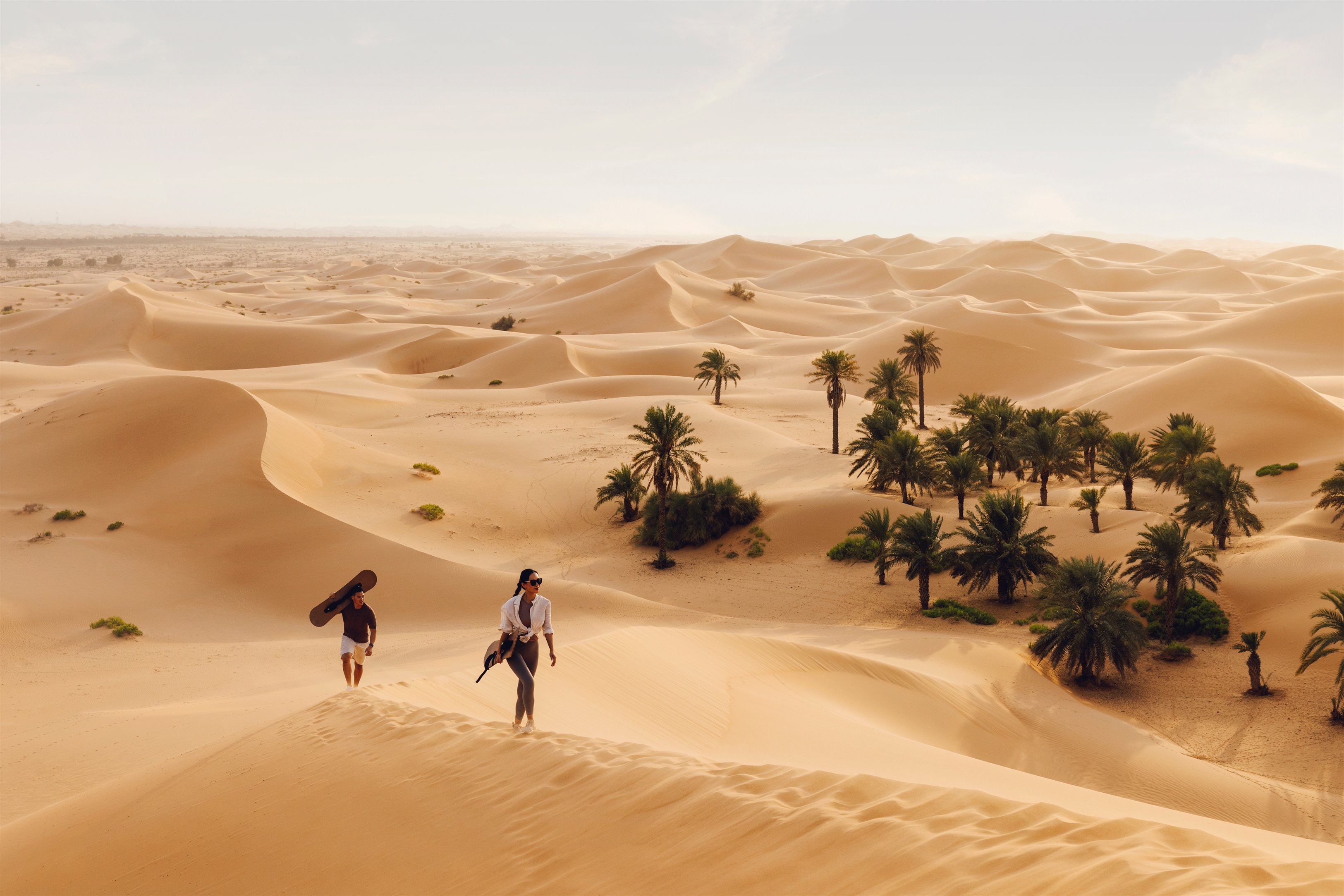 Two people trekking through golden sand dunes near a desert oasis in Abu Dhabi.