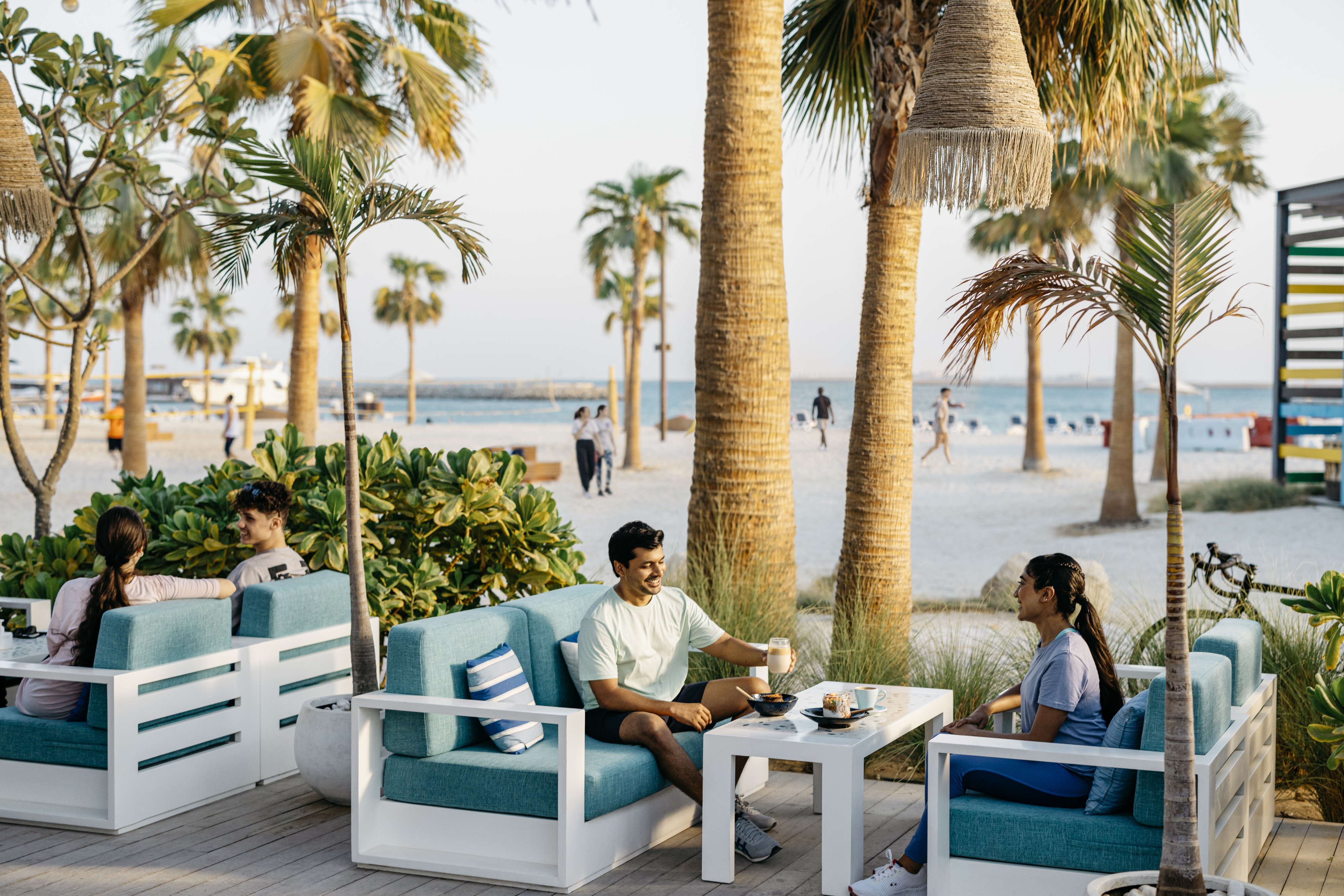  Sur l'île de Hudayriyat, des amis profitent d'un moment convivial autour d'un café, entourés par des palmiers et la mer scintillante.